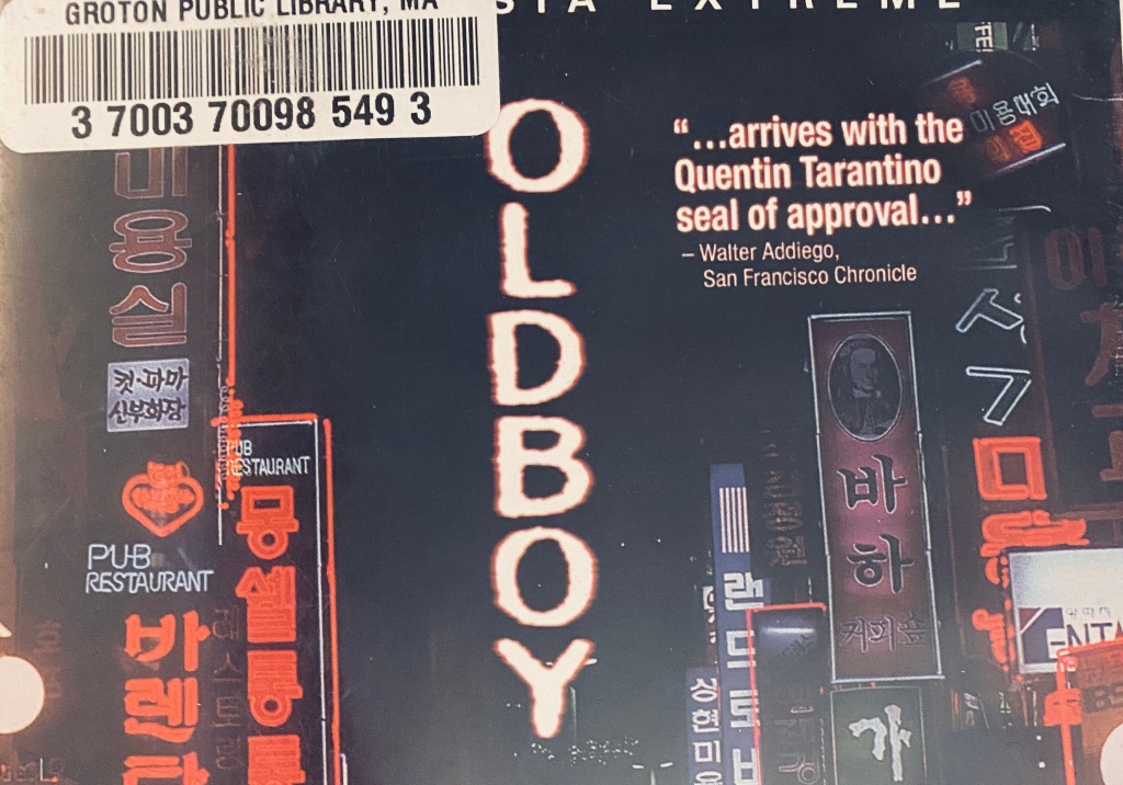 A closeup of he DVD case for Oldboy: the title is in glowing letters against a night sky, surrounded by neon signs in Korean. A sticker reading "Groton Public Library" is on the upper left corner, and the following quote is on the upper right: "...arrives with the Quentin Tarantino seal of approval" -- Walter Addiego, San Francisco Chronicle